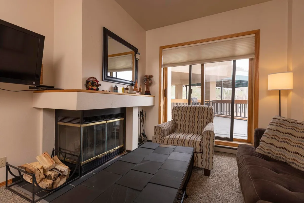 This image centers on a warm and inviting living room with a wood-burning fireplace as the focal point. Comfortable seating is arranged to encourage relaxation and conversation, while a large window lets in natural light and offers glimpses of the serene mountain surroundings. The overall atmosphere is cozy and perfect for unwinding after a day outdoors.