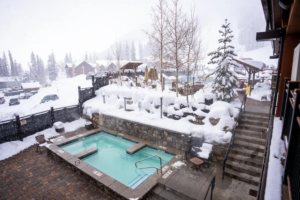 This image captures a steaming hot tub set against a snowy landscape, surrounded by snow-covered trees and nearby resort buildings. The serene winter setting suggests relaxation and tranquility, highlighting the hot tub as a luxurious amenity perfect for soothing tired muscles after a day of adventure.