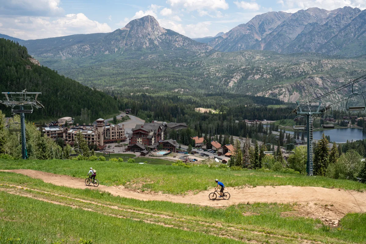 A broad exterior view featuring a stunning mountain range, a tranquil lake, resort buildings, a ski lift, and winding bike trails. The energetic scene captures a variety of outdoor activities and communal spaces where couples can explore and relax together.