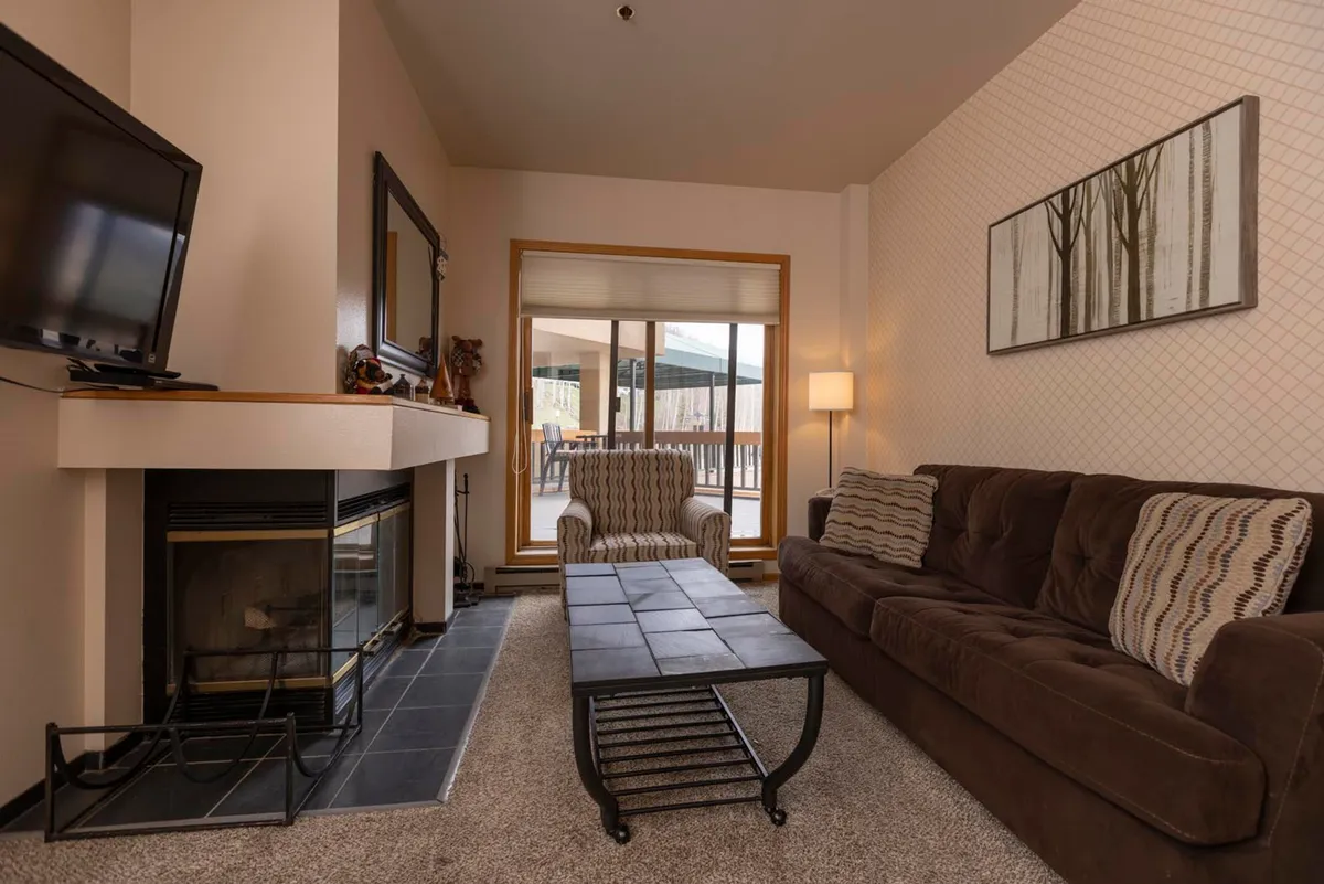 The image showcases a warm, inviting living area with a glowing wood-burning fireplace as the centerpiece, a plush sofa perfect for lounging, a coffee table, and large windows that let in natural light and hint at mountain views. Artwork on the walls adds a touch of mountain charm, and the overall setting is cozy and welcoming.