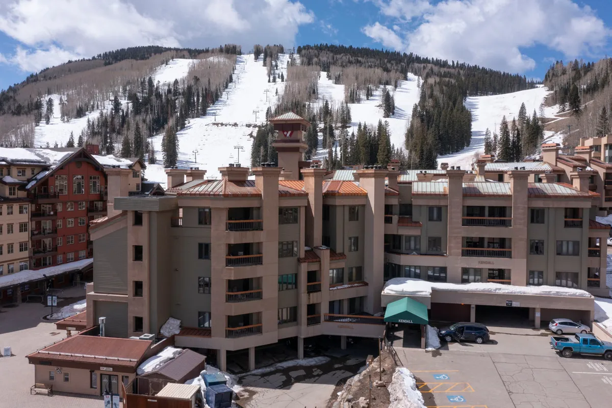 This image shows the exterior of the resort building with the parking area in the foreground and majestic mountains rising in the background. The building facade is inviting, and the parking lot highlights the convenience of on-site garage parking for guests.