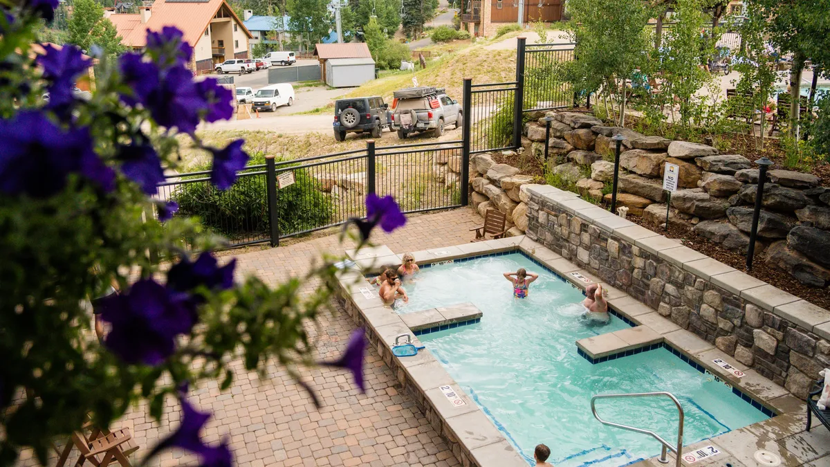 The photo displays a sparkling outdoor swimming pool and a hot tub, framed by blooming flowers and a scenic landscape. The setting exudes a cheerful, resort-like ambiance and invites thoughts of socializing, swimming, and lounging in the sun.