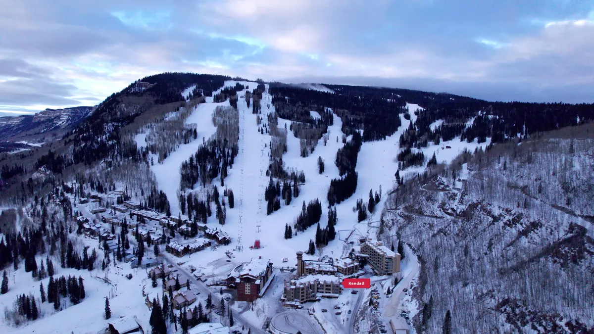 This image showcases an inviting mountain scene with pristine ski slopes winding down toward a cozy lodging complex, all framed by towering peaks and ski lifts. The early morning light casts a warm glow, suggesting both the adventure of the day ahead and the tranquil comfort awaiting couples as they return to their accommodations.